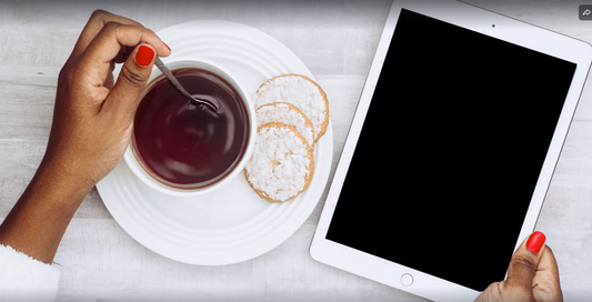 A person holds a spoon in a cup of tea and an iPad, with a plate of cookies. 